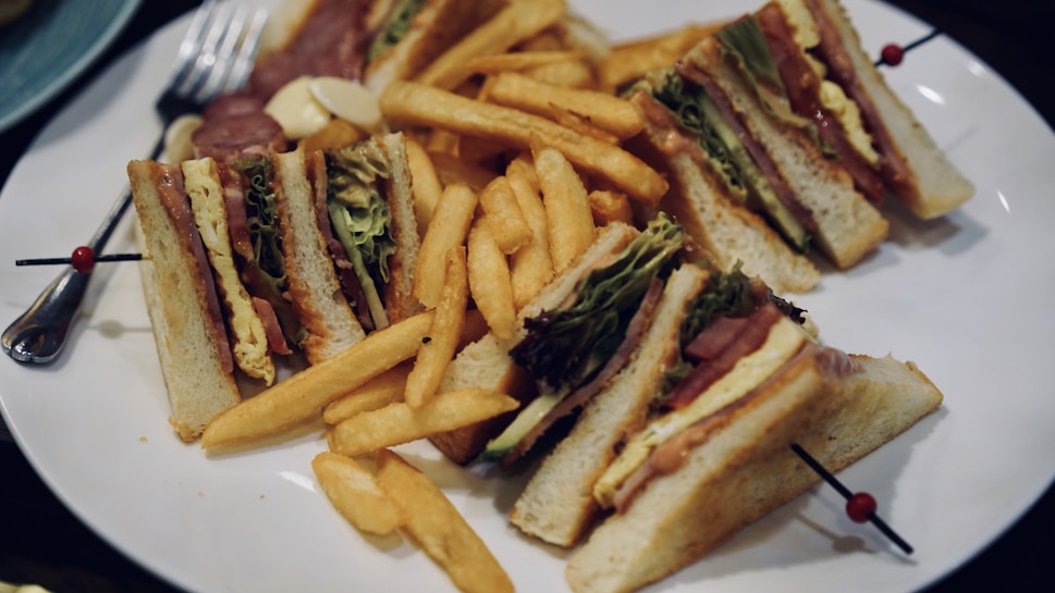 A plate filled with club sandwiches and french fries. The sandwiches include layers of lettuce, tomato, cheese, and various deli meats, skewered to hold them together. The fries are golden and crispy, scattered around the sandwiches.