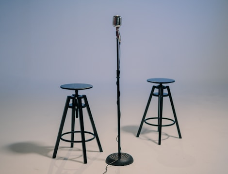 A vintage-style microphone is positioned on a stand at the center of the image, flanked by two tall black stools. The background is plain and softly lit, emphasizing the objects in the foreground.