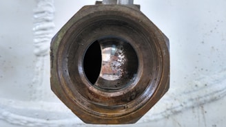 A close-up view of a rusty, metallic pipe with a hexagonal nut. The interior of the pipe shows signs of wear and oxidation, and there is a reflective surface inside. The background is out of focus, with a light gray or off-white color.