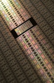 A close-up view of a semiconductor wafer, featuring a grid pattern of many microchips. The surface reflects light in various iridescent colors, highlighting intricate electronic circuits and layers. The image emphasizes precision engineering and technological complexity.