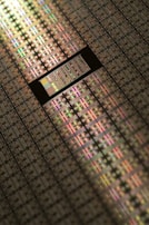 A close-up view of a semiconductor wafer, featuring a grid pattern of many microchips. The surface reflects light in various iridescent colors, highlighting intricate electronic circuits and layers. The image emphasizes precision engineering and technological complexity.