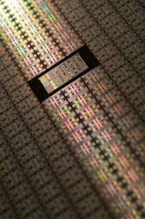 A close-up view of a semiconductor wafer, featuring a grid pattern of many microchips. The surface reflects light in various iridescent colors, highlighting intricate electronic circuits and layers. The image emphasizes precision engineering and technological complexity.