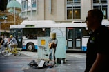 A street performer dressed in an eclectic outfit with a bucket covering their head plays an electric guitar. Nearby, a bicycle rider wearing a yellow helmet rides by, and a large city bus is visible in the background. Several pedestrians are walking, and an individual in a dark shirt is in the foreground.
