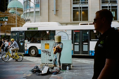 A street performer dressed in an eclectic outfit with a bucket covering their head plays an electric guitar. Nearby, a bicycle rider wearing a yellow helmet rides by, and a large city bus is visible in the background. Several pedestrians are walking, and an individual in a dark shirt is in the foreground.