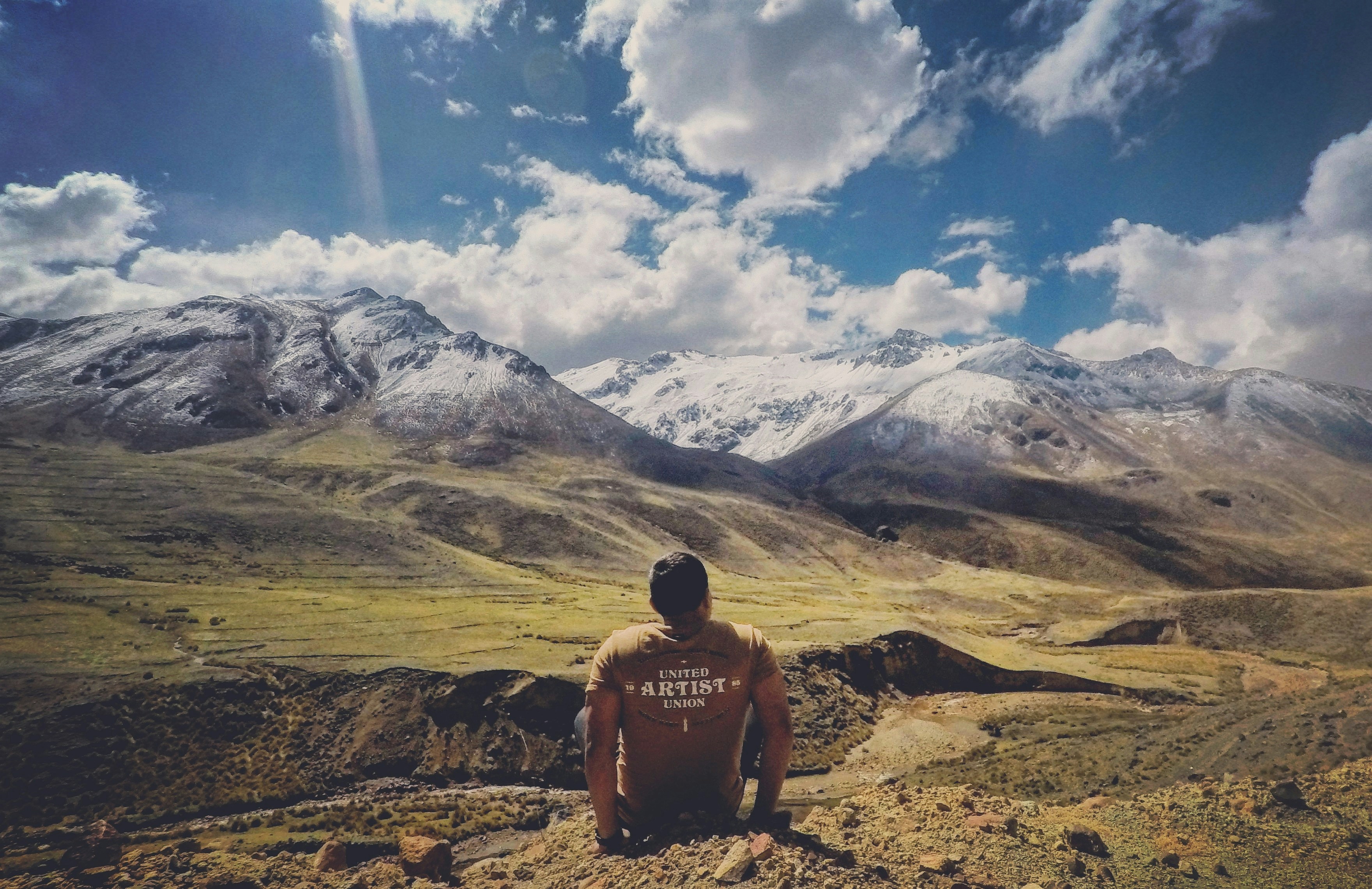 man in black tank top sitting on brown rock mountain under blue sky and white clouds
