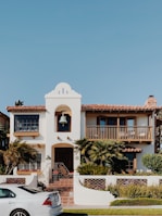 A two-story house with a Spanish architectural style has a white exterior, a red-tiled roof, and a prominent bell tower above the entrance. The front of the house features a balcony with wooden railings, palm trees and other shrubs in the garden, and a paved walkway leading to the wooden front door. A white car is parked on the street in front of the house. The sky is clear and blue.