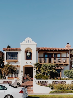 A two-story house with a Spanish architectural style has a white exterior, a red-tiled roof, and a prominent bell tower above the entrance. The front of the house features a balcony with wooden railings, palm trees and other shrubs in the garden, and a paved walkway leading to the wooden front door. A white car is parked on the street in front of the house. The sky is clear and blue.