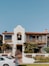 A two-story house with a Spanish architectural style has a white exterior, a red-tiled roof, and a prominent bell tower above the entrance. The front of the house features a balcony with wooden railings, palm trees and other shrubs in the garden, and a paved walkway leading to the wooden front door. A white car is parked on the street in front of the house. The sky is clear and blue.