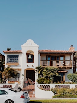 A two-story house with a Spanish architectural style has a white exterior, a red-tiled roof, and a prominent bell tower above the entrance. The front of the house features a balcony with wooden railings, palm trees and other shrubs in the garden, and a paved walkway leading to the wooden front door. A white car is parked on the street in front of the house. The sky is clear and blue.