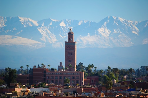 A tall, intricate minaret rises above a cityscape with palm trees, set against a backdrop of majestic snow-capped mountains under a clear blue sky. The architecture reflects traditional Islamic design, contributing to a serene and historic ambiance.