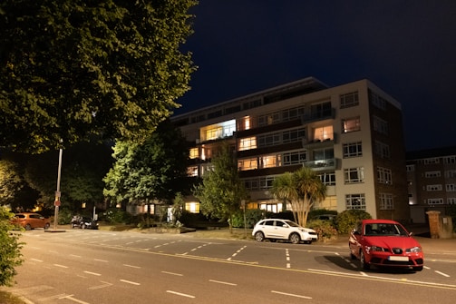A multi-story residential building is illuminated by warm lighting from several windows. In front of the building, there are parked cars in designated parking spaces along a quiet street. The scene is set at night, and the area is surrounded by trees with dense foliage.