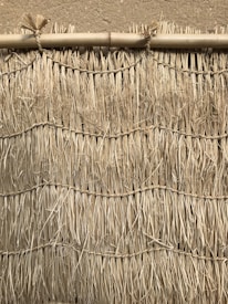 A close-up view of a traditional, textured wall created from tightly bound straw or reed bundles, reinforced with horizontal bamboo. The surface features rough, dry fibers with natural variations in color and texture.