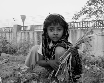 A young child with a solemn expression is crouched down outdoors, holding a bundle of dried twigs. The background features a concrete wall topped with barbed wire and sparse vegetation.