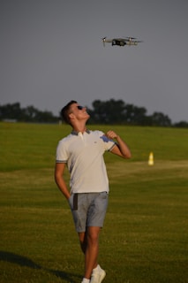 A person wearing a white polo shirt and blue shorts is standing on a grassy field, looking up at a hovering drone. The sky is clear, and there are trees and a yellow cone in the background.
