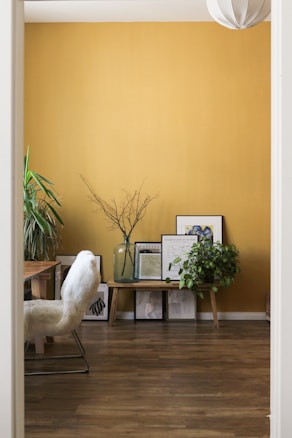 A cozy room features a mustard yellow wall adorned with framed art prints resting on a low wooden bench. A large glass vase with twigs and a lush green plant add a touch of nature. A white, fluffy chair sits next to a wooden table, and a modern pendant light hangs from the ceiling.