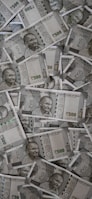 A large pile of Indian currency notes, featuring the portrait of Mahatma Gandhi. The notes are primarily 500 denomination, arranged in a slightly disordered manner.