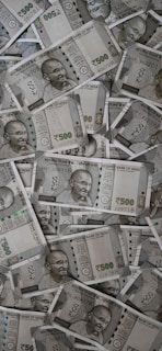 A large pile of Indian currency notes, featuring the portrait of Mahatma Gandhi. The notes are primarily 500 denomination, arranged in a slightly disordered manner.