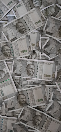 A large pile of Indian currency notes, featuring the portrait of Mahatma Gandhi. The notes are primarily 500 denomination, arranged in a slightly disordered manner.