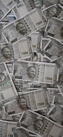 A large pile of Indian currency notes, featuring the portrait of Mahatma Gandhi. The notes are primarily 500 denomination, arranged in a slightly disordered manner.