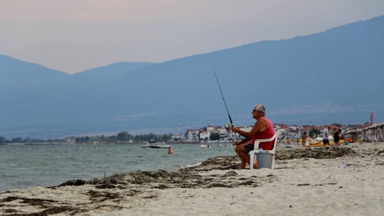 man in red shirt sitting on white and blue boat on beach during daytime in Paralia Greece