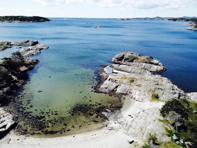 An aerial view of a coastal landscape featuring clear blue waters, rocky shores, and a small bay with sandy beaches. There is a white cabin situated on a rock outcrop near the water. Several small islands and additional rocky formations are visible in the distance, under a partly cloudy sky.