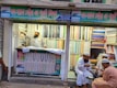 A textile shop displays an array of colorful fabrics neatly stacked on shelves. A man in traditional attire appears to be organizing or handling the fabrics. Three other men in similar attire are seated outside the shop, engaged in conversation while having tea. The shop has signs with text in what appears to be Bengali or a similar script.