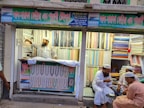 A textile shop displays an array of colorful fabrics neatly stacked on shelves. A man in traditional attire appears to be organizing or handling the fabrics. Three other men in similar attire are seated outside the shop, engaged in conversation while having tea. The shop has signs with text in what appears to be Bengali or a similar script.