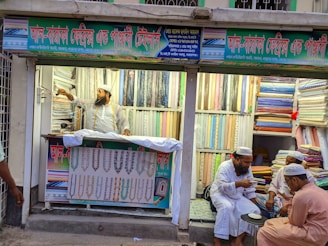 A textile shop displays an array of colorful fabrics neatly stacked on shelves. A man in traditional attire appears to be organizing or handling the fabrics. Three other men in similar attire are seated outside the shop, engaged in conversation while having tea. The shop has signs with text in what appears to be Bengali or a similar script.