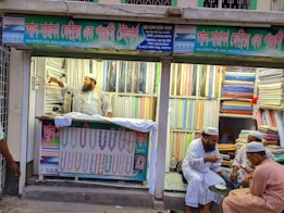 A textile shop displays an array of colorful fabrics neatly stacked on shelves. A man in traditional attire appears to be organizing or handling the fabrics. Three other men in similar attire are seated outside the shop, engaged in conversation while having tea. The shop has signs with text in what appears to be Bengali or a similar script.