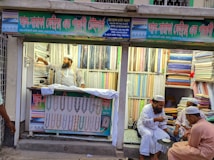 A textile shop displays an array of colorful fabrics neatly stacked on shelves. A man in traditional attire appears to be organizing or handling the fabrics. Three other men in similar attire are seated outside the shop, engaged in conversation while having tea. The shop has signs with text in what appears to be Bengali or a similar script.