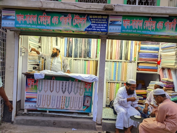A textile shop displays an array of colorful fabrics neatly stacked on shelves. A man in traditional attire appears to be organizing or handling the fabrics. Three other men in similar attire are seated outside the shop, engaged in conversation while having tea. The shop has signs with text in what appears to be Bengali or a similar script.