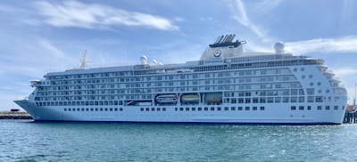 A large, white luxury cruise ship docked at a harbor, with multiple decks and rows of windows. The name 'The World' is visible on the side of the ship. The sky is clear with wispy clouds, and the water is calm.