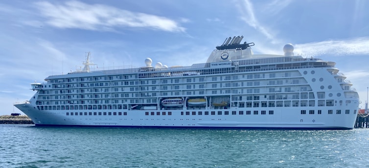A large, white luxury cruise ship docked at a harbor, with multiple decks and rows of windows. The name 'The World' is visible on the side of the ship. The sky is clear with wispy clouds, and the water is calm.
