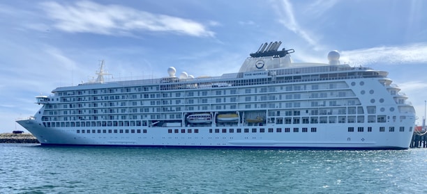 A large, white luxury cruise ship docked at a harbor, with multiple decks and rows of windows. The name 'The World' is visible on the side of the ship. The sky is clear with wispy clouds, and the water is calm.