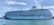 A large, white luxury cruise ship docked at a harbor, with multiple decks and rows of windows. The name 'The World' is visible on the side of the ship. The sky is clear with wispy clouds, and the water is calm.
