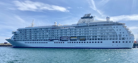 A large, white luxury cruise ship docked at a harbor, with multiple decks and rows of windows. The name 'The World' is visible on the side of the ship. The sky is clear with wispy clouds, and the water is calm.