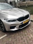 A silver BMW car is parked on a cobblestone street next to a body of water, displaying a yellow license plate with an NL marking. The car features prominent front grilles and sleek headlights.