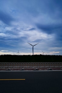 Wind turbines stand tall against a cloudy sky, set behind a dark silhouette of trees. A paved road with a yellow line runs parallel to a stone wall at the foreground.