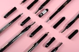 Several makeup brushes and eyebrow pencils are neatly arranged on a pink background. The items are in various states of assembly, with some showing bristles and others revealing the pencil tips.