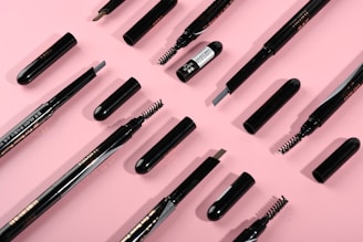 Several makeup brushes and eyebrow pencils are neatly arranged on a pink background. The items are in various states of assembly, with some showing bristles and others revealing the pencil tips.