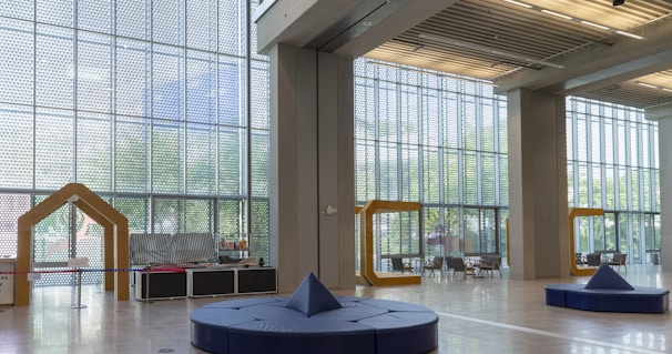A spacious and well-lit interior with large floor-to-ceiling windows featuring a geometric design. The room contains modern seating arrangements and decorative structures in the shape of frame-like yellow arches. Light wooden floors and ceiling panels add to the contemporary feel.