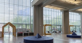 A spacious and well-lit interior with large floor-to-ceiling windows featuring a geometric design. The room contains modern seating arrangements and decorative structures in the shape of frame-like yellow arches. Light wooden floors and ceiling panels add to the contemporary feel.