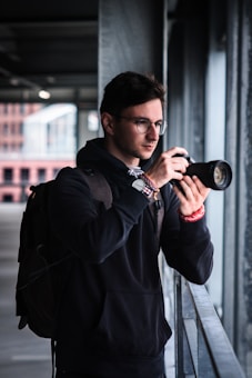 A person with glasses is holding a camera, preparing to take a photograph while standing in an urban setting. They are wearing a dark hoodie and carrying a backpack, suggesting they might be exploring or on a photographic assignment. The background shows an industrial or modern building environment, partially blurred.