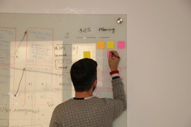 A person is writing on a transparent whiteboard with colorful sticky notes. Various diagrams and text are visible on the board, likely related to planning or brainstorming. The person is slightly turned away from the camera and is holding a marker.