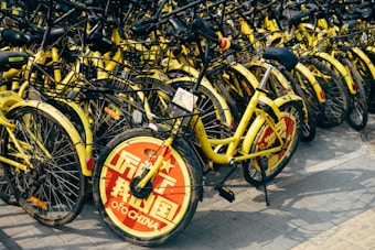 A large cluster of yellow rental bicycles with black handlebars is tightly packed together. The front wheel of each bike is covered with a red circular plate displaying white text, with some writing visible. They are parked on a paved surface.