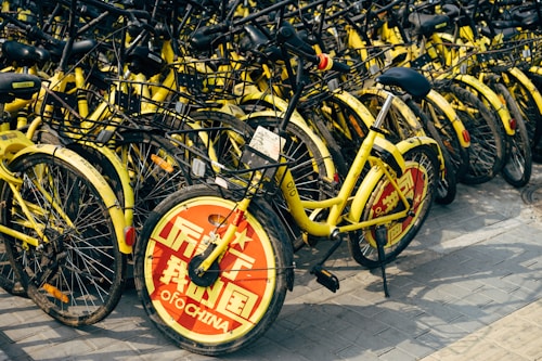 A large cluster of yellow rental bicycles with black handlebars is tightly packed together. The front wheel of each bike is covered with a red circular plate displaying white text, with some writing visible. They are parked on a paved surface.