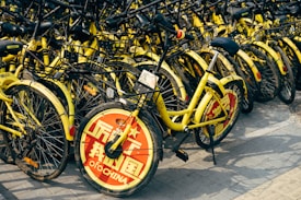 A large cluster of yellow rental bicycles with black handlebars is tightly packed together. The front wheel of each bike is covered with a red circular plate displaying white text, with some writing visible. They are parked on a paved surface.