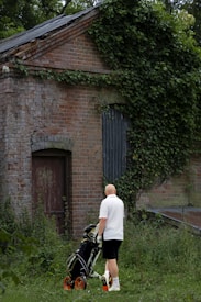 A man wearing a white shirt and black shorts stands in front of an old brick building overgrown with ivy and vegetation. He is pushing a golf cart with bright orange wheels. The building appears to be abandoned and surrounded by greenery.
