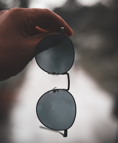 A hand holds a pair of round sunglasses with dark lenses against a blurry outdoor background, primarily in muted tones.