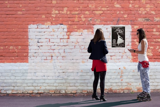Two women stand in front of a red and white brick wall. One is dressed in a navy jacket and red skirt, while the other wears patterned pants and a white top with rollerblades. They both face a poster on the wall with information about a ballet performance.